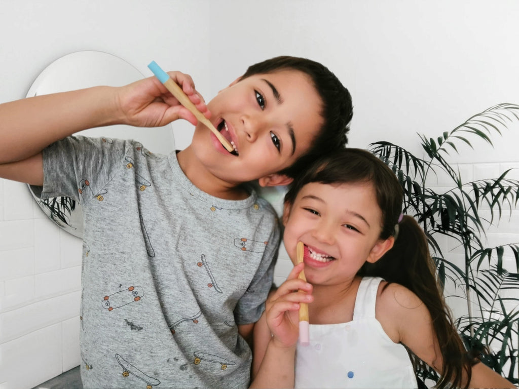 A boy is holding a blue bamboo toothbrush and a girl is holding a pink bamboo toothbrush